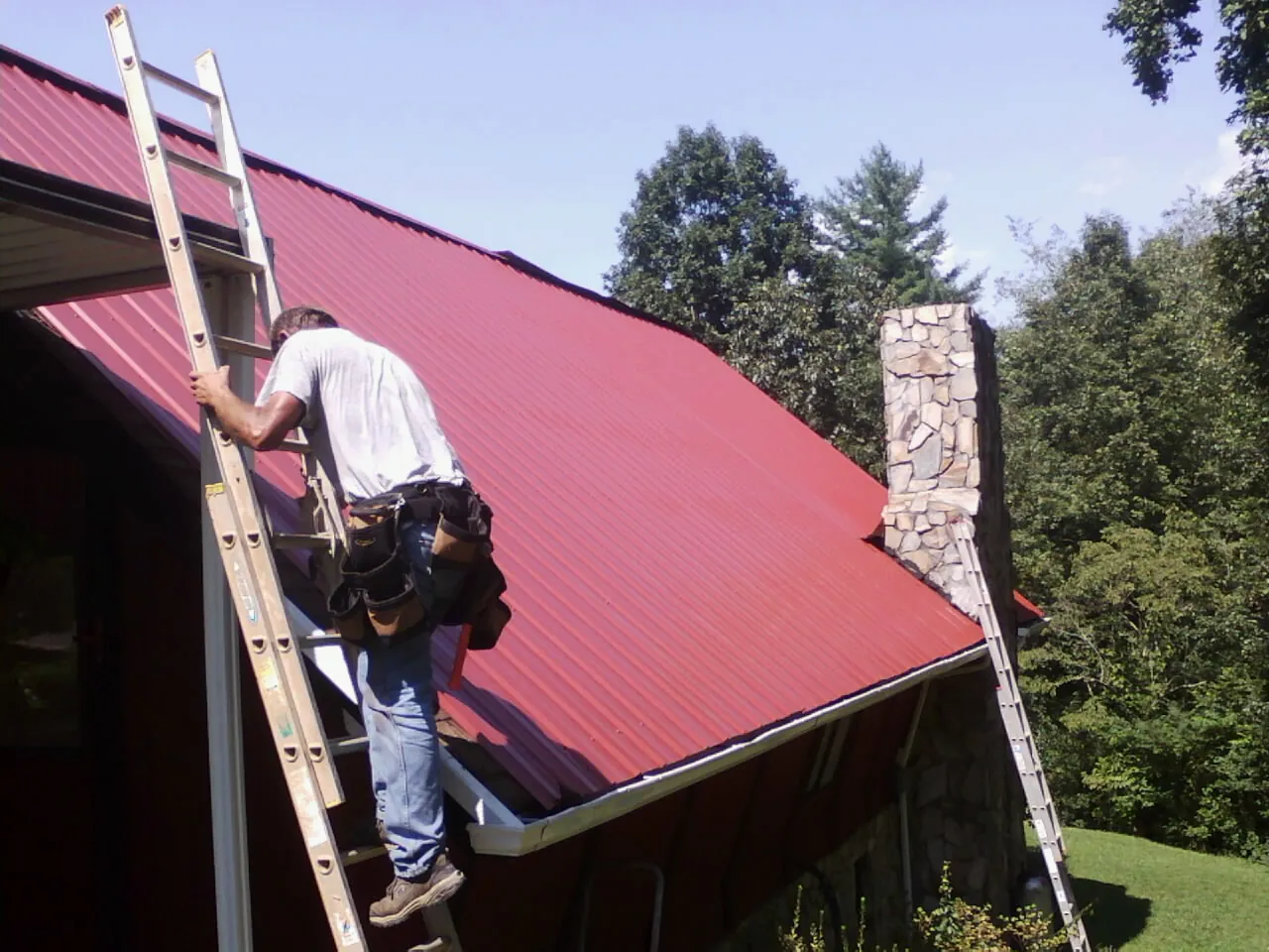 Roofer on a ladder beside a red metal roof and stone chimney — Soffit & Fascia Repair by Roofing Brothers in Amazonia